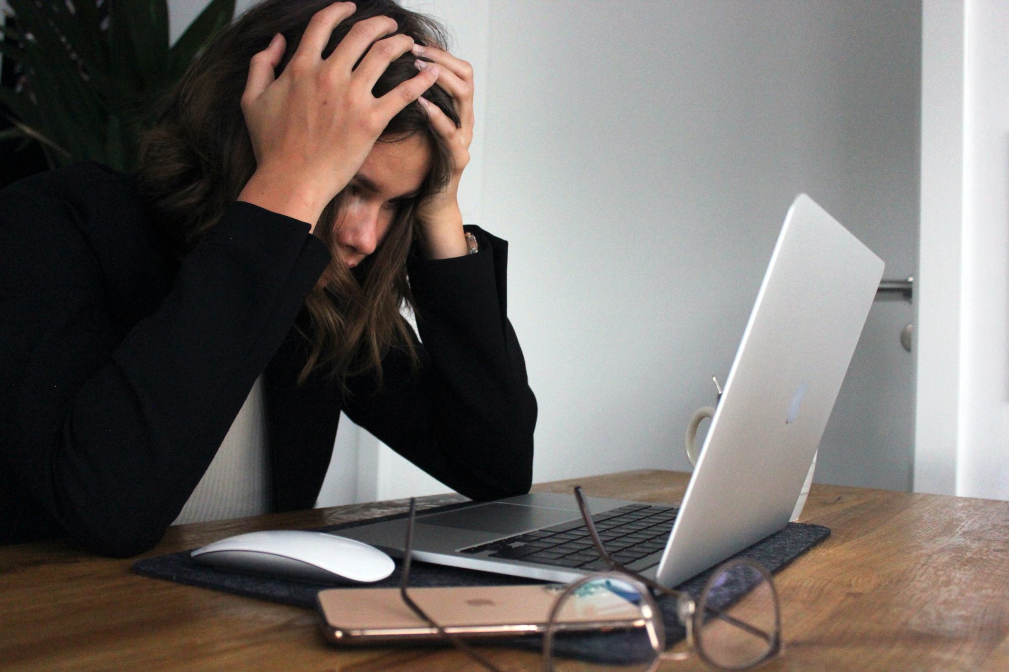 Woman sat at desk holding her head in her hands looking stressed with IT equipment in front of her.